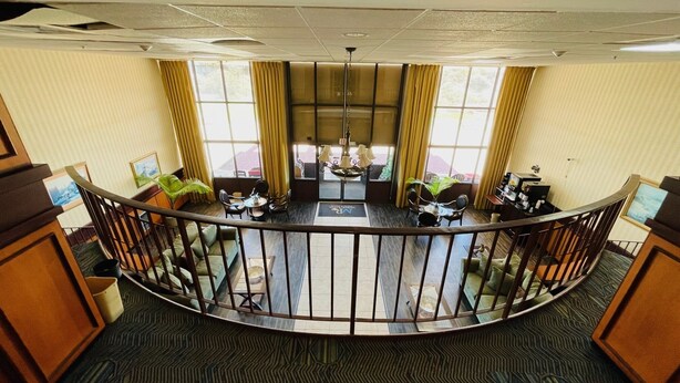 A hotel lobby with a curved railing, a central seating area with tables and chairs, and large windows with curtains.