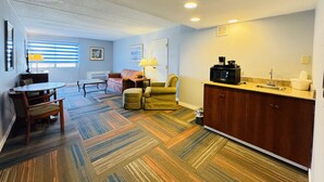 A compact hotel room with a kitchenette, a dining area with a table and chairs, a sofa, and a patterned carpet.