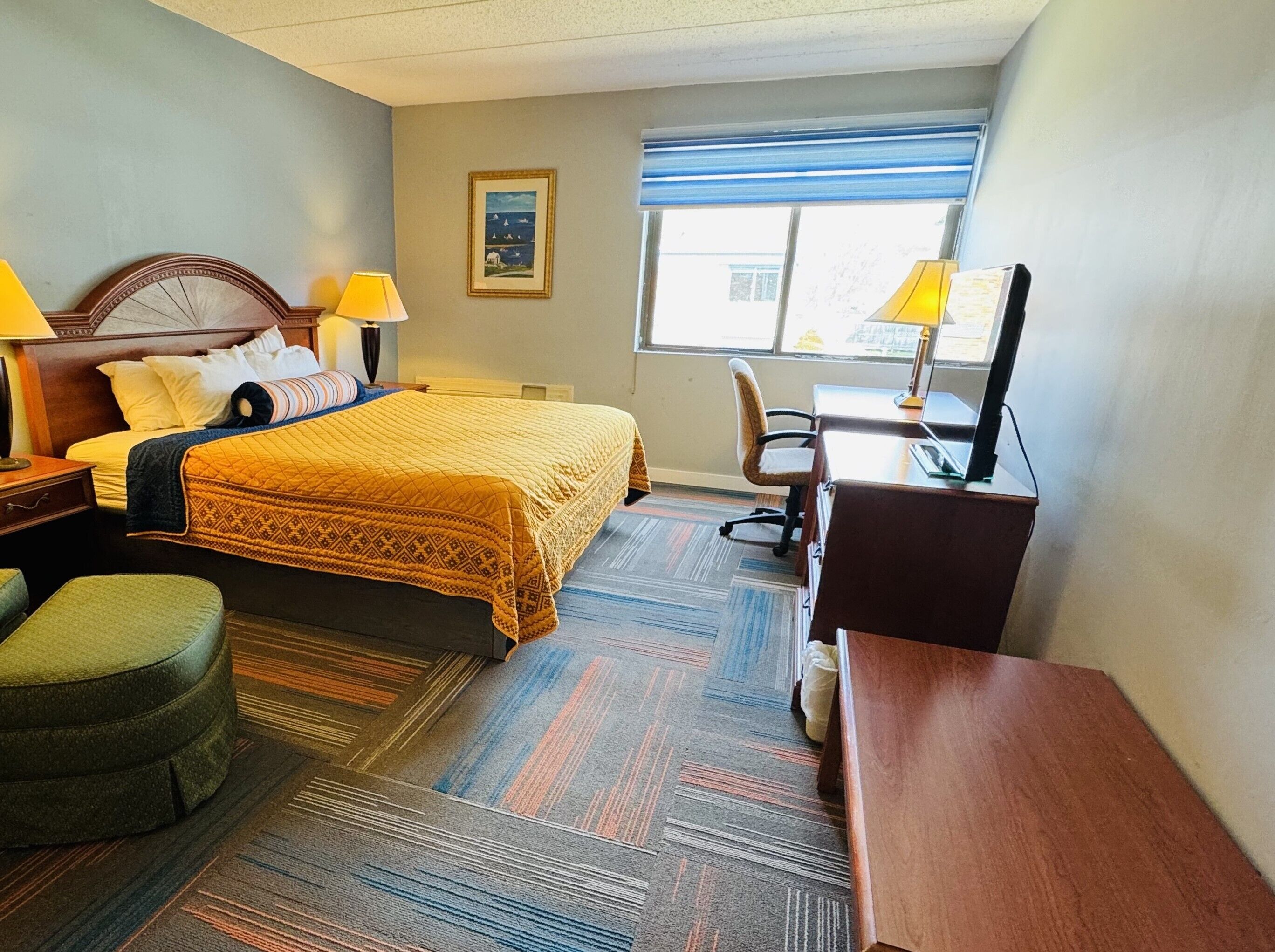A hotel room with a bed, two lamps, a desk, a chair, a green ottoman, and a window with blinds.