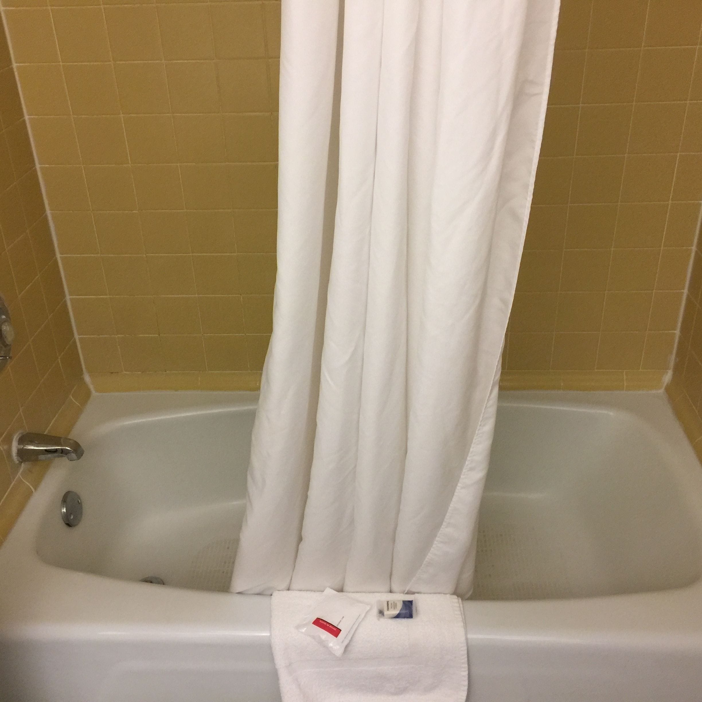 Una bañera con una cortina blanca, una toalla blanca y una pequeña botella de champú en el borde.