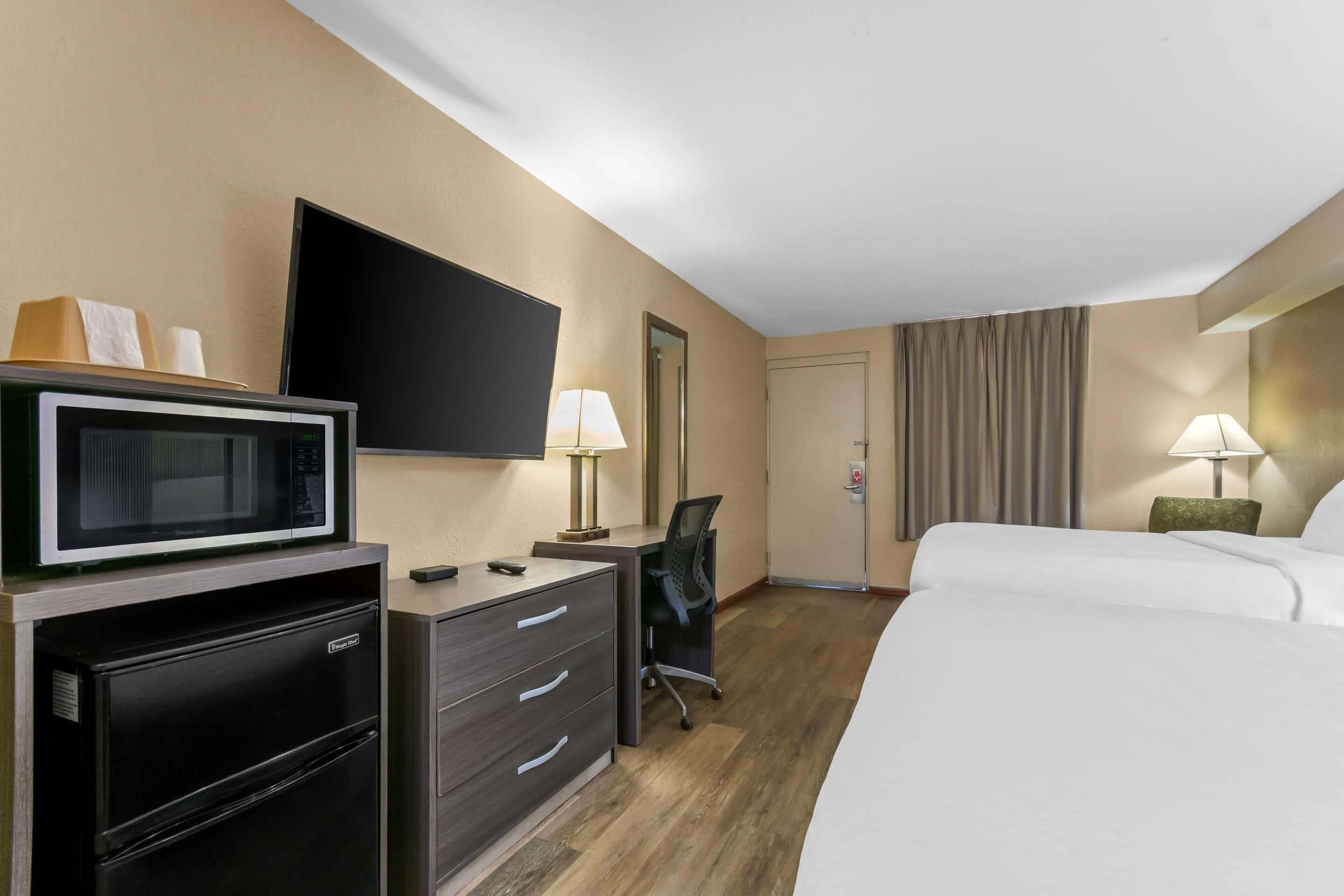 A hotel room with a bed, a desk with a computer, a microwave, a refrigerator, a flat-screen TV, and two lamps.