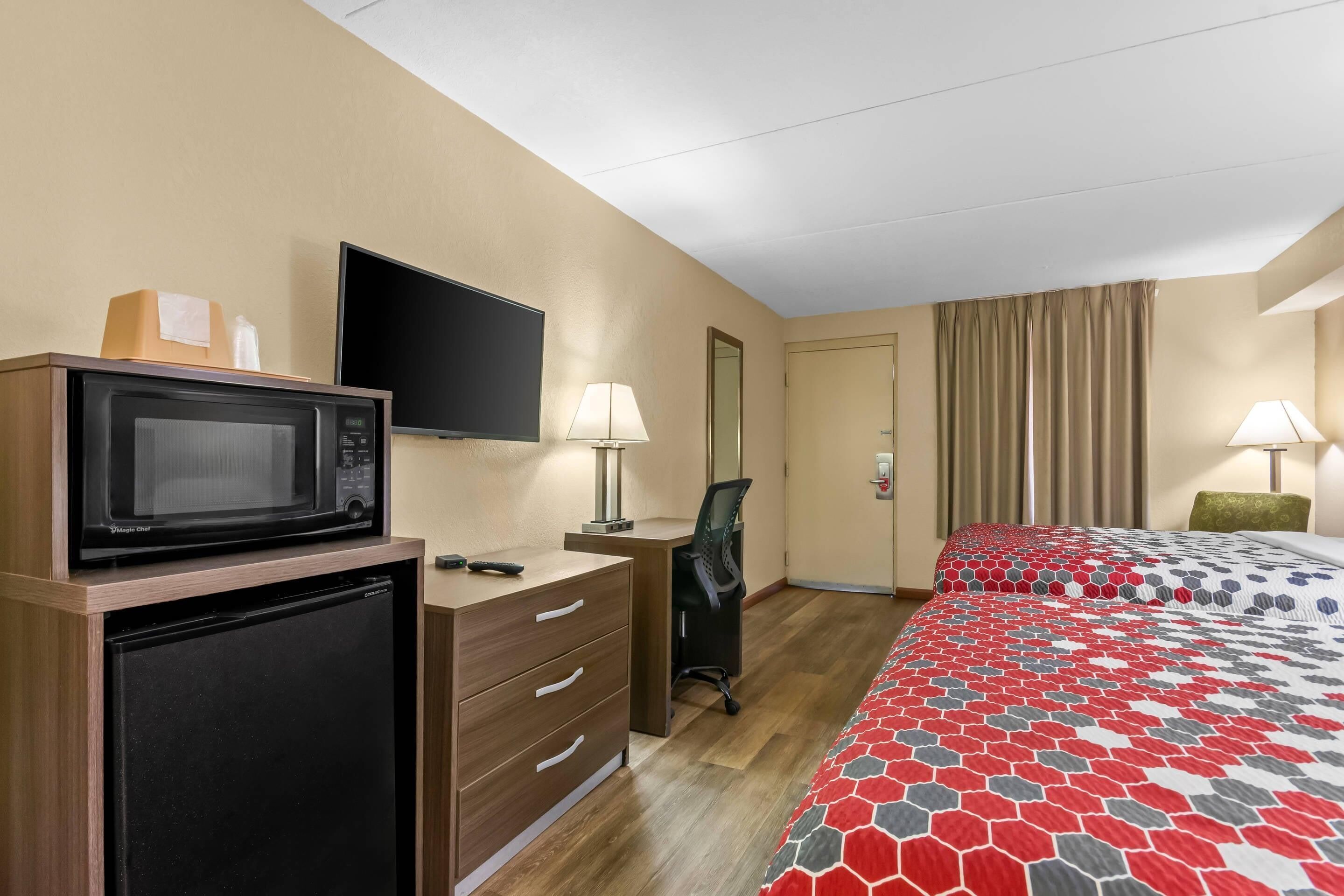 A hotel room with a microwave, TV, desk, chair, bed, and curtains.