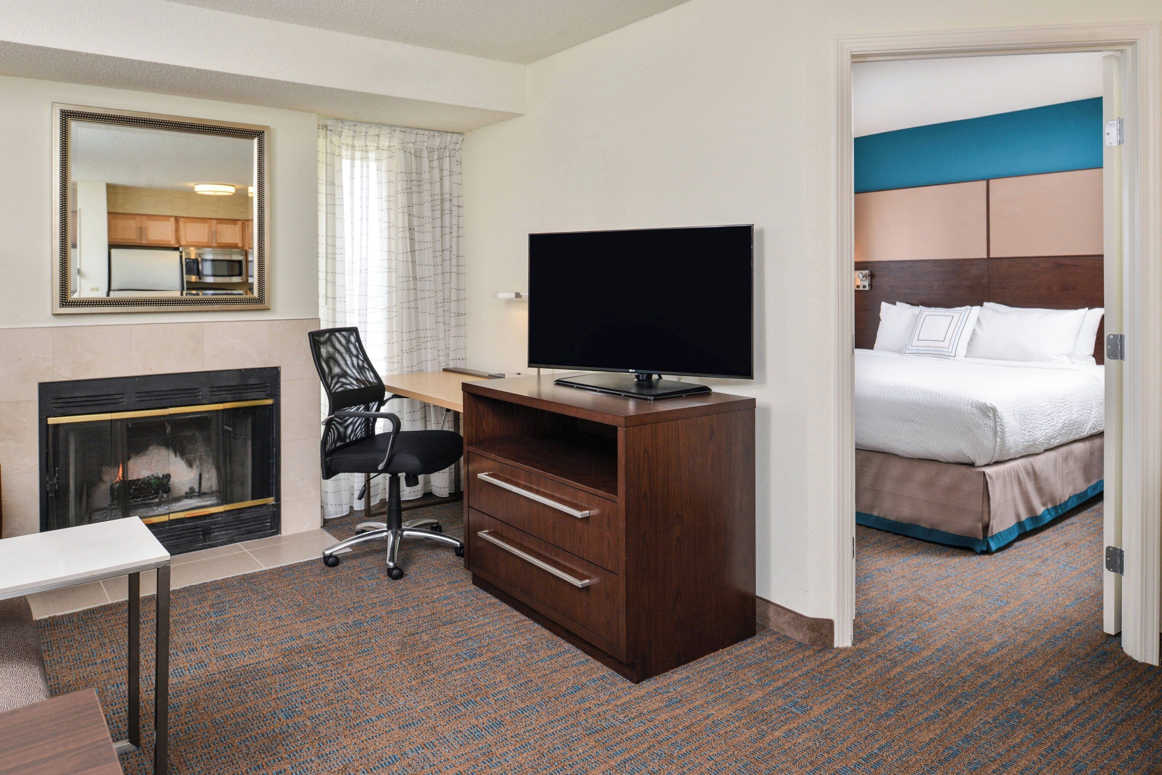 A hotel room with a fireplace, a desk with a flat-screen TV, a bed with a blue headboard, and a large mirror on the wall.