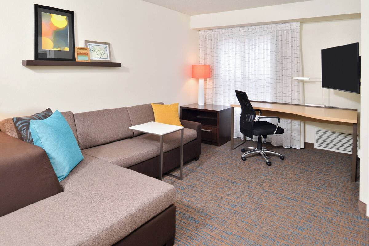 A modern hotel room with a sofa, a small table, a desk with a chair, a TV, and a window with curtains.