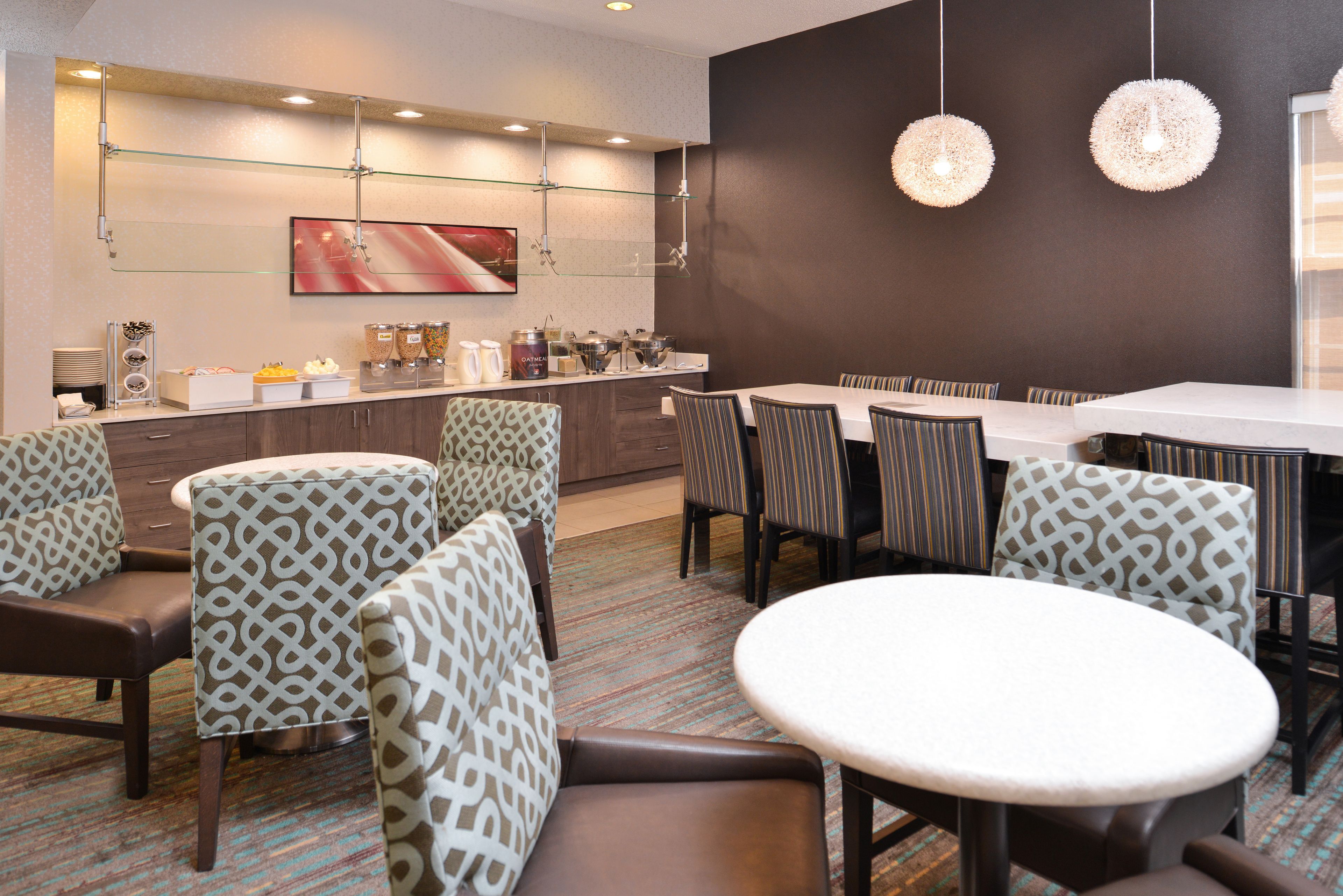 Habitación de hotel moderna con zona de comedor que presenta sillas estampadas y una mesa redonda, una barra de buffet con alimentos, y un cuadro abstracto grande en la pared.