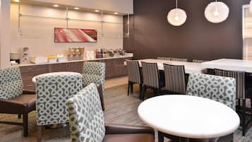 Habitación de hotel moderna con zona de comedor que presenta sillas estampadas y una mesa redonda, una barra de buffet con alimentos, y un cuadro abstracto grande en la pared.