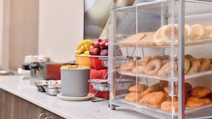 Un expositor de pastelería con diversos productos horneados, un frutero y una máquina de café sobre una encimera.