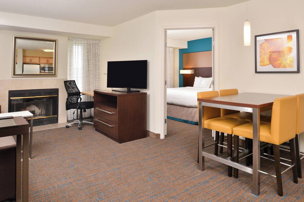 A hotel room with a TV, a desk, a chair, a bed, and a dining area with yellow chairs.