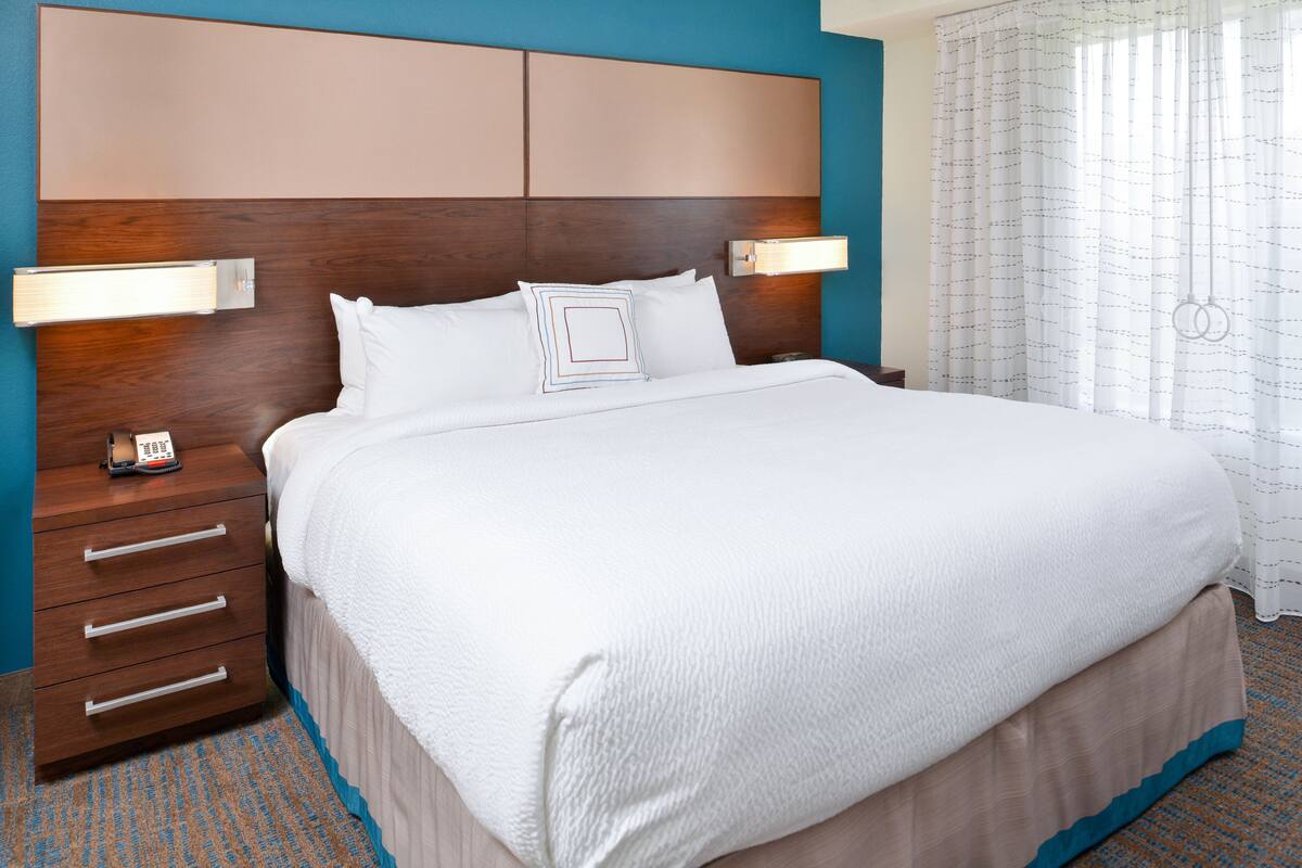 A neatly made bed with a wooden headboard, a bedside table with a phone, and a patterned carpet.