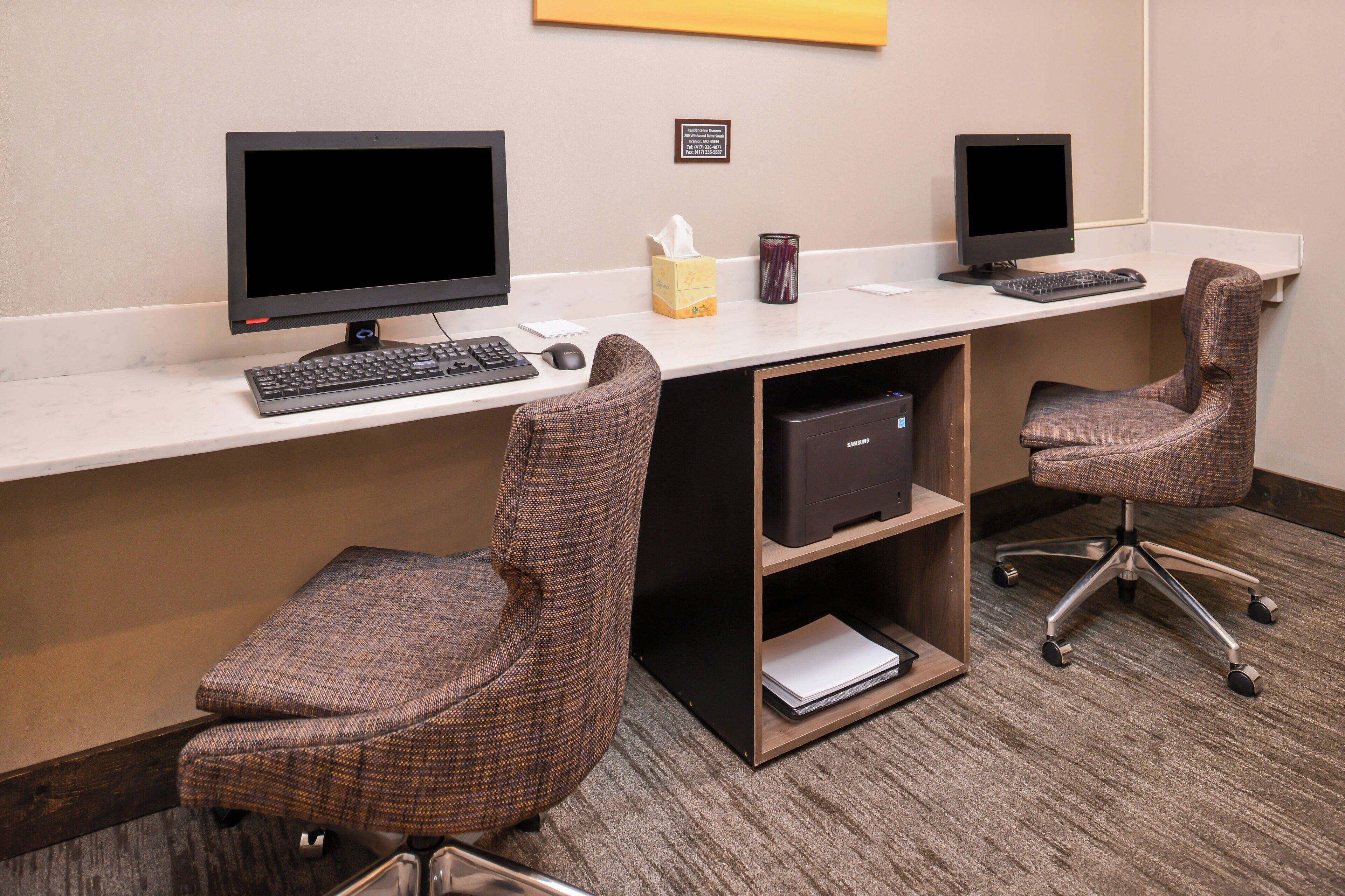 A hotel workspace with two computer desks, each with a monitor, keyboard, and mouse. There are two upholstered office chairs. A tissue box and a mug are on the desks. A small shelf with a printer is under one desk.
