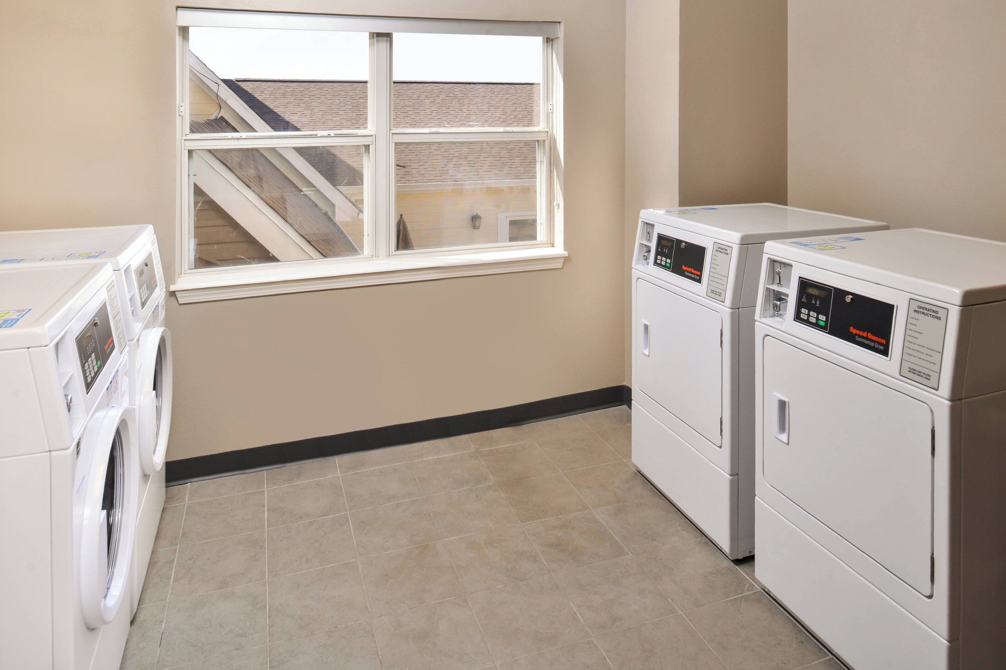 A laundry room with two front-loading washers and dryers, a window with a view of a building, and a tiled floor.