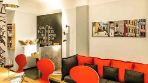 A modern lounge area with a wooden table, red and black seating, a chalkboard with text, and a cityscape painting. - ibis Nuernberg Altstadt (Nuremberg)