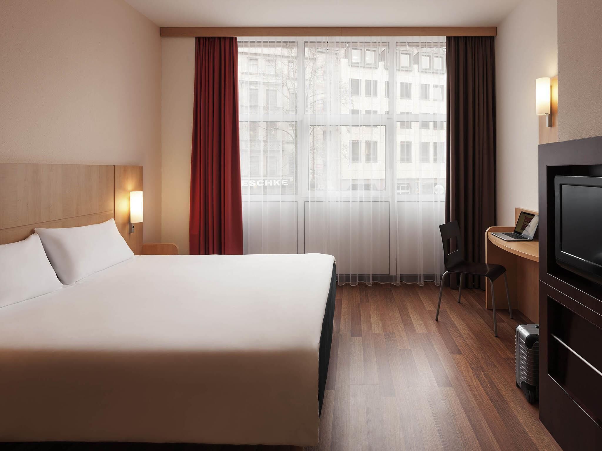 A hotel room with a large bed, a desk with a laptop, a television, and a view of buildings through the window.