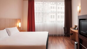 Quarto de hotel com uma cama, uma escrivaninha com um laptop, uma televisão e uma janela com cortinas.