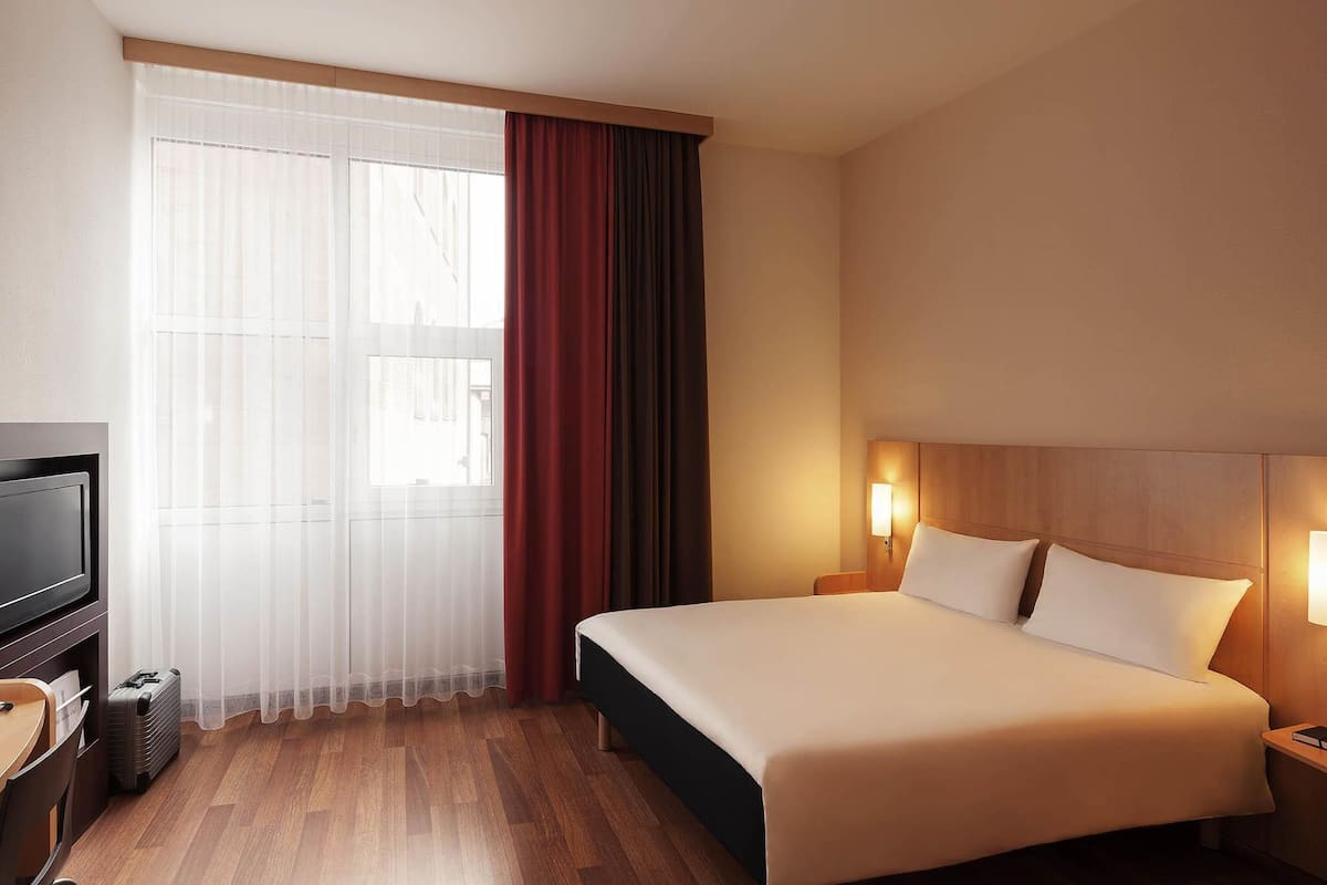 Una habitación de hotel con cama, lámparas de noche, televisión y ventana con cortinas.