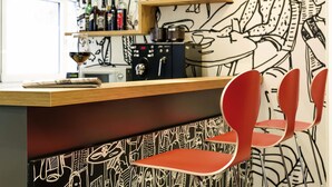 Uma área de bar com balcão, prateleiras com garrafas, copos e um mural.