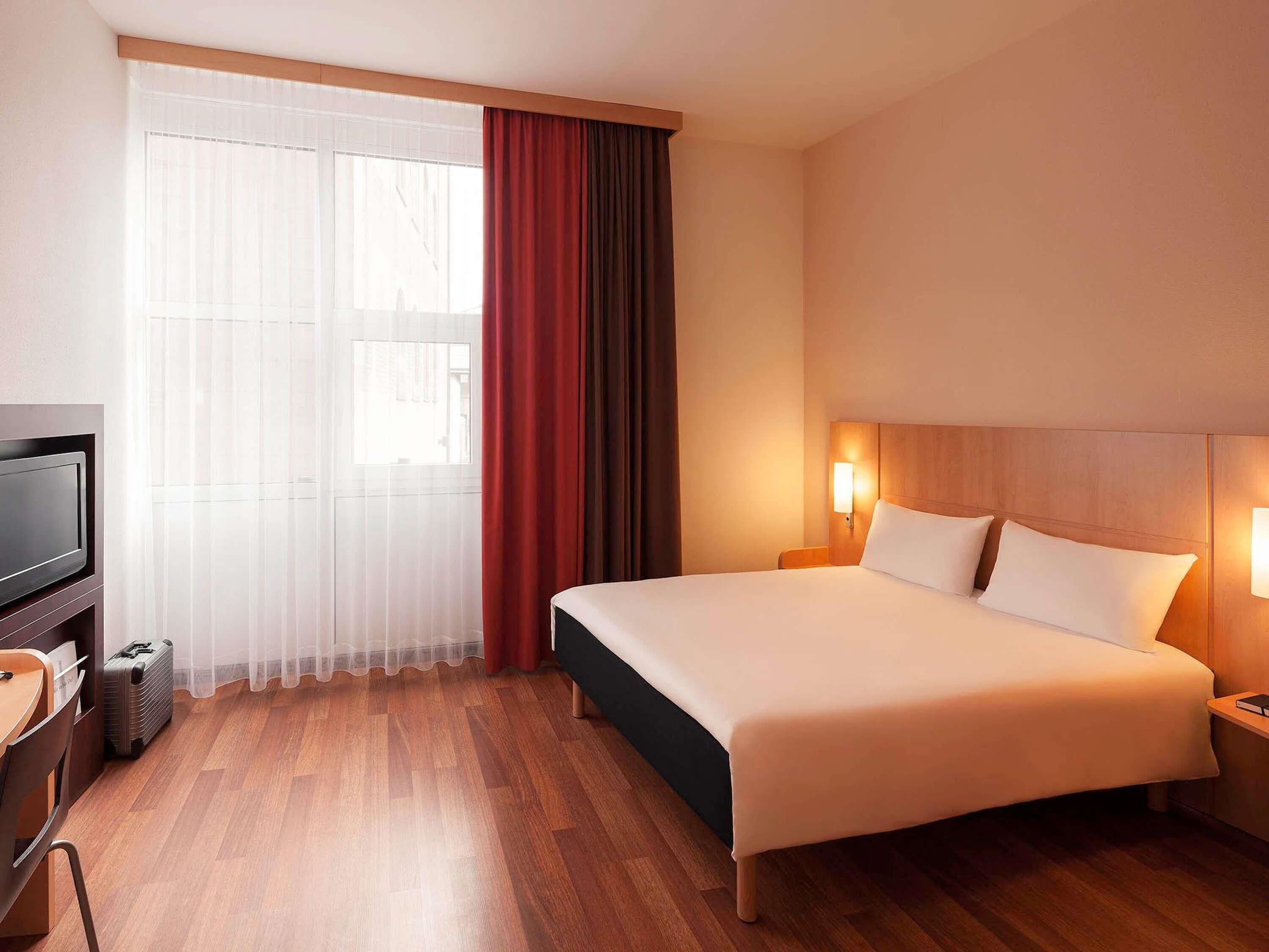 A hotel room with a bed, a TV, a desk, and a window with curtains.