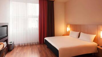 A hotel room with a bed, a TV, a desk, and a window with curtains.