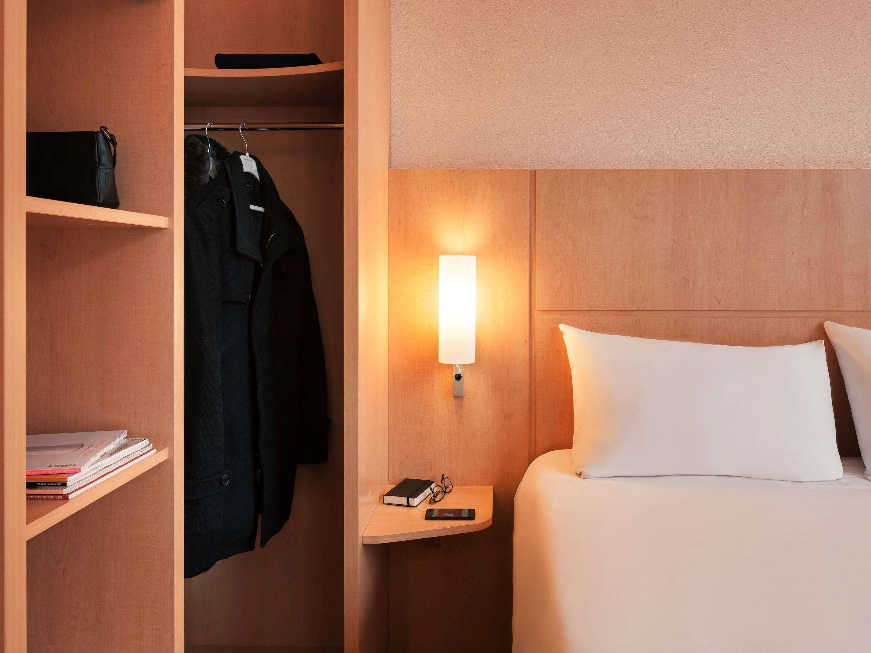 Habitación de hotel con cama, mesita de noche con lámpara, armario y estante con libros.