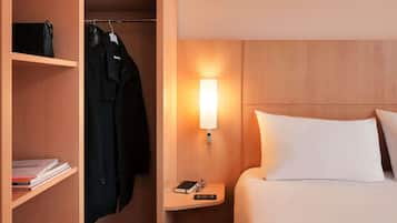 Habitación de hotel con cama, mesita de noche con lámpara, armario y estante con libros.