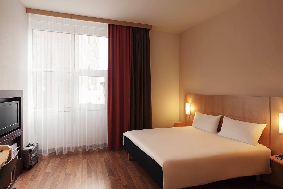 A hotel room with a bed, bedside lamps, a TV, and a window with curtains.
