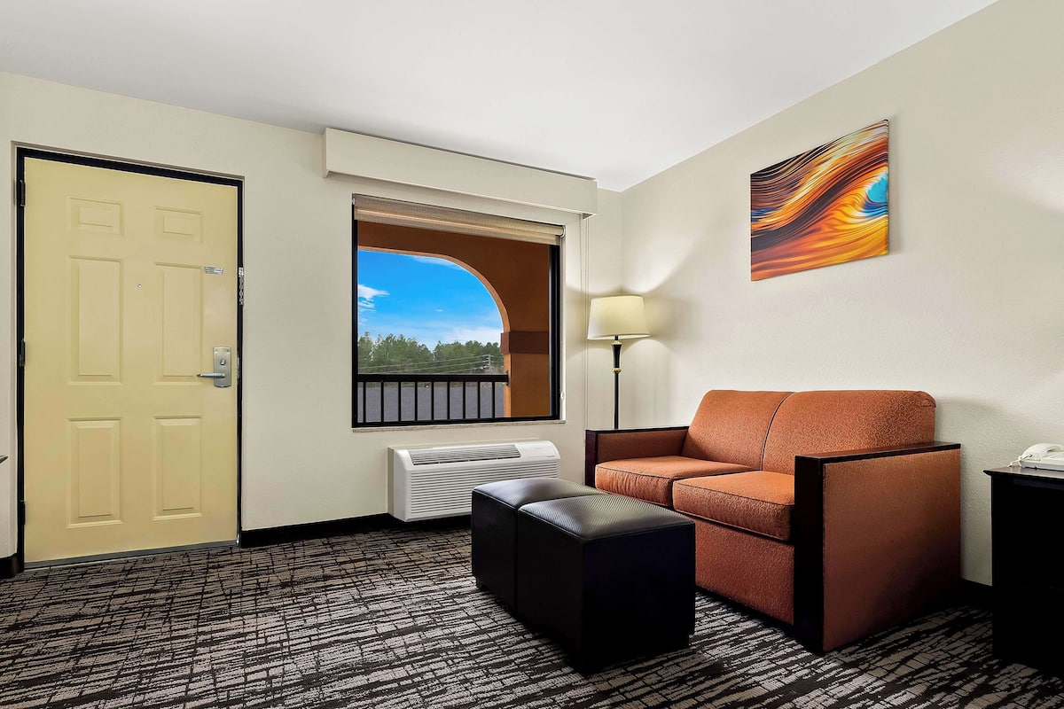 A hotel room with a beige door, a window with a view of trees, a brown sofa, a black ottoman, and a floor lamp.