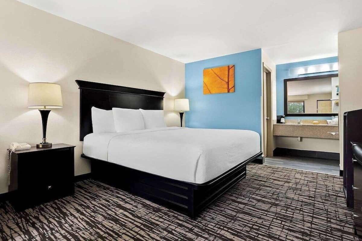 A hotel room with a bed, two bedside lamps, a nightstand, a blue accent wall, a bathroom with a sink and mirror, and a patterned carpet.