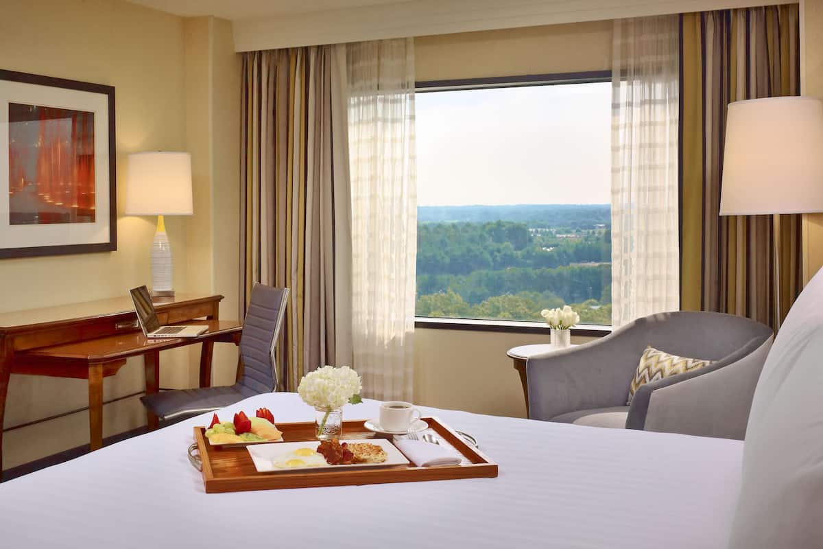 Une chambre d'hôtel avec un lit, un bureau, une chaise, une petite table sur laquelle se trouve un plateau de petit-déjeuner, et une fenêtre donnant sur des arbres.