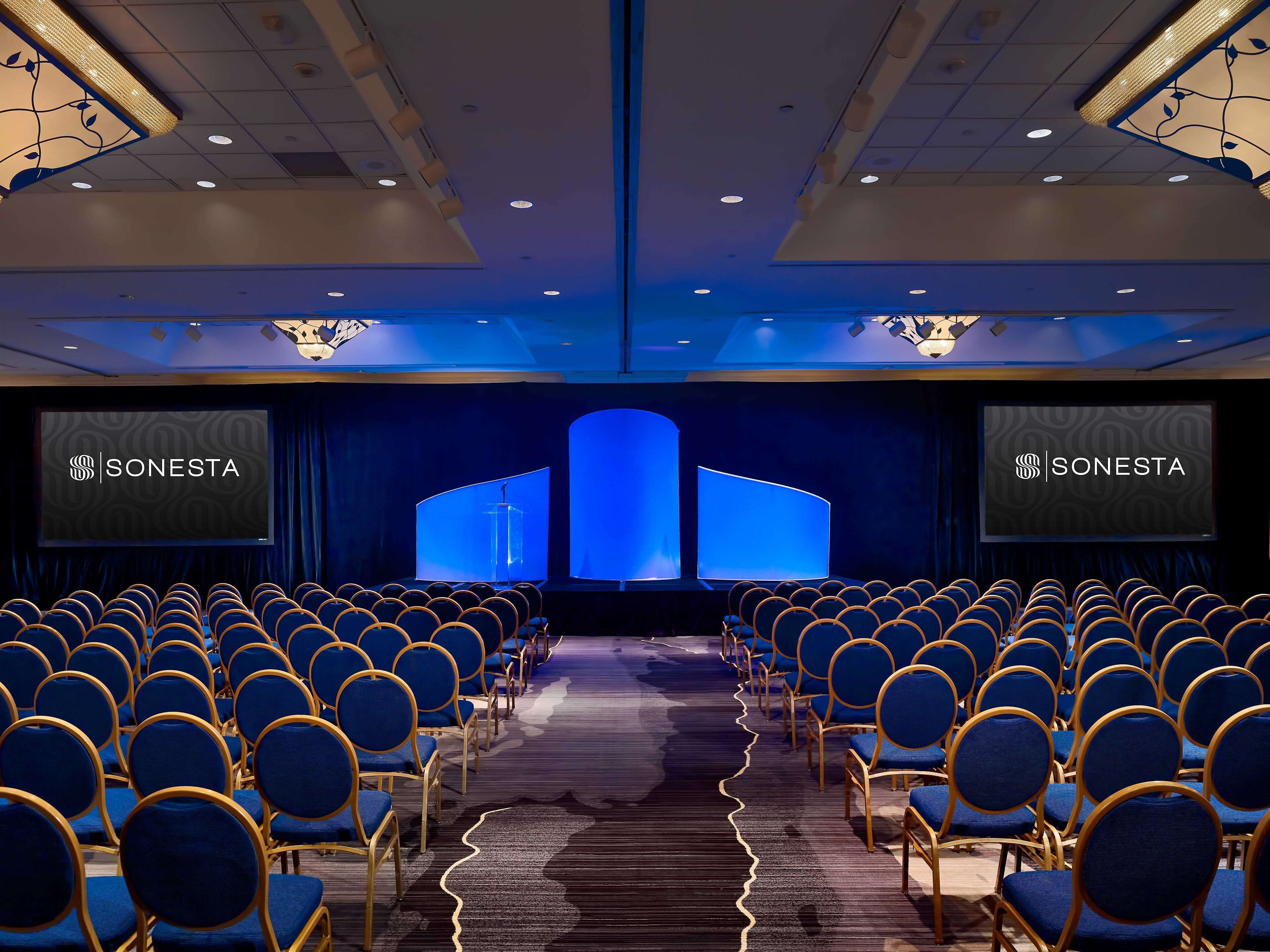 Un salón de conferencias con filas de sillas azules, un escenario con un atril y dos pantallas que muestran el logotipo de SONESTA.