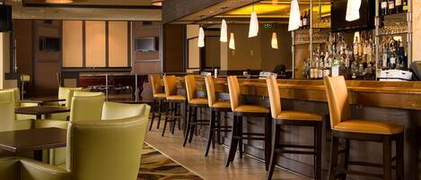 A bar area with multiple seating options including bar stools and chairs, a bar counter with a variety of bottles and glasses, and overhead lighting fixtures.