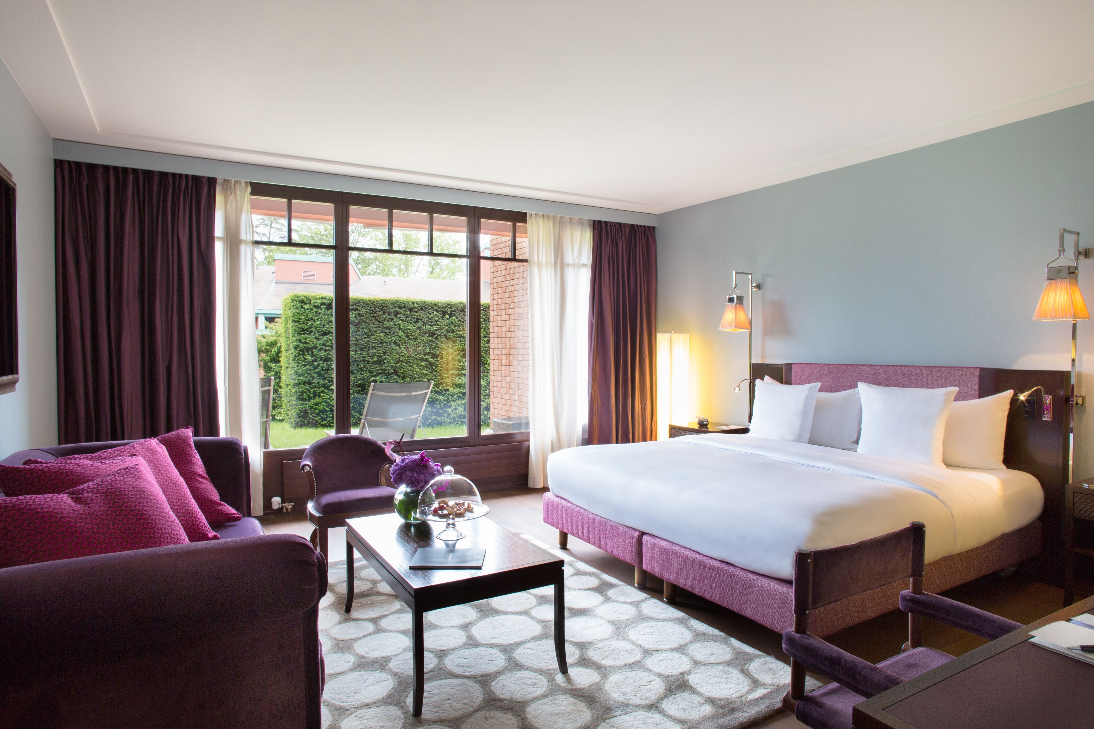 A hotel room with a large bed, a sofa, a coffee table, and a view of the outside.
