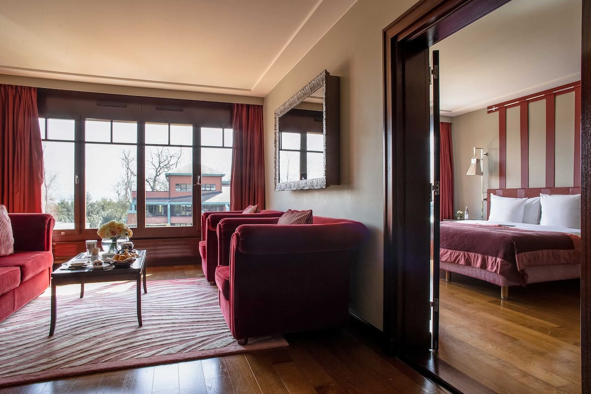 A hotel room with a large window, a red sofa, a bed with a red bedspread, and a mirror on the wall.