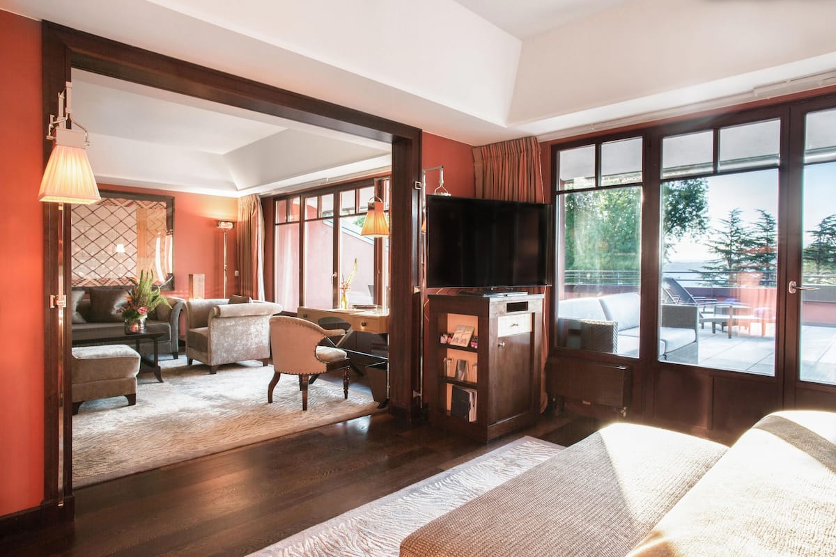 A hotel room with a sofa, armchairs, a TV, and a large window offering a view of the outdoors.