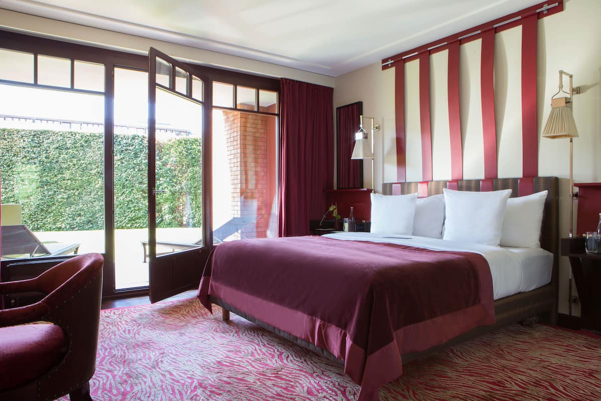A hotel room with a large bed, a desk, and a chair. There is a window with a view of greenery outside.