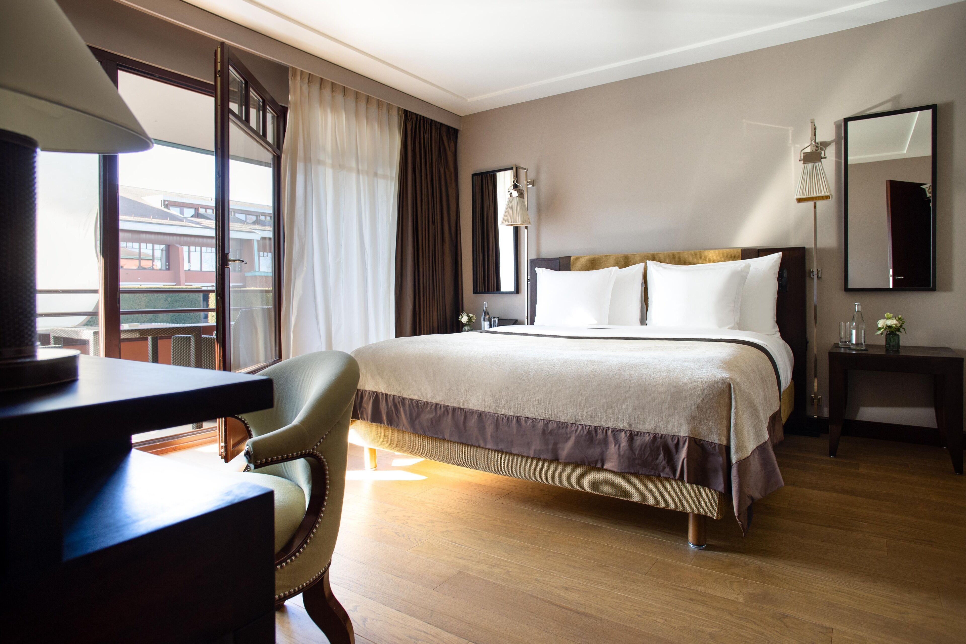 A hotel room with a large bed, a desk, a chair, a bedside table, a lamp, a mirror, and a view of buildings through the window.