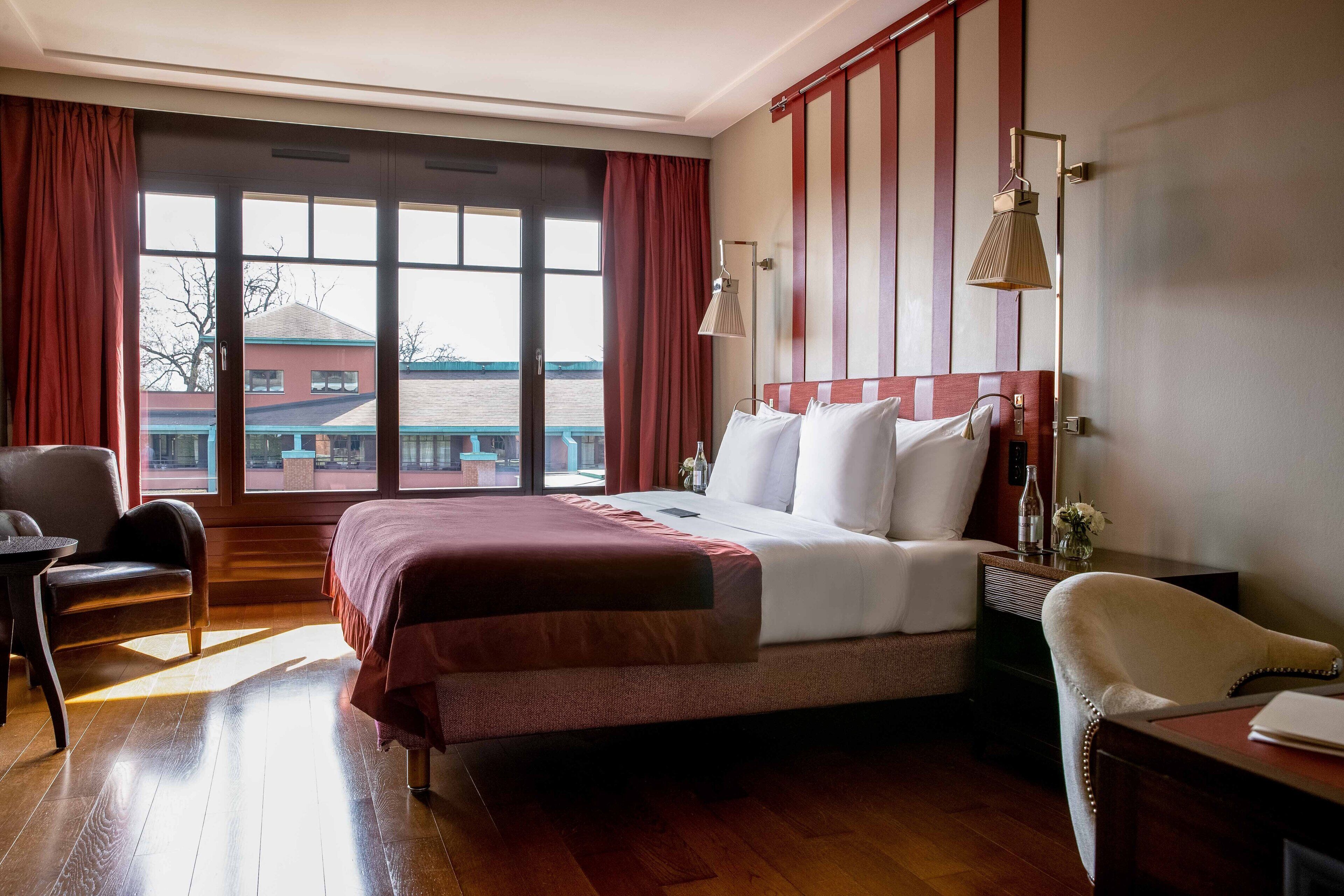 A hotel room with a large bed, a desk, a chair, and a view of a building outside.