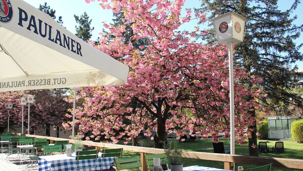 An outdoor seating area with tables covered in checkered tablecloths, a sign reading 'PAULANER,' and a clock post. - Holiday Inn Munich - South by IHG (Munich)