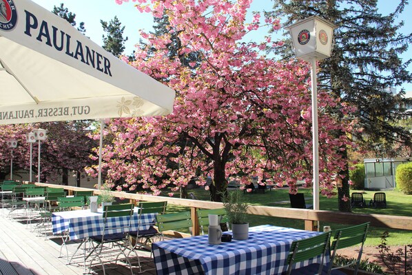 An outdoor seating area with tables covered in checkered tablecloths, a sign reading 'PAULANER,' and a clock post. - Holiday Inn Munich - South by IHG (Munich)