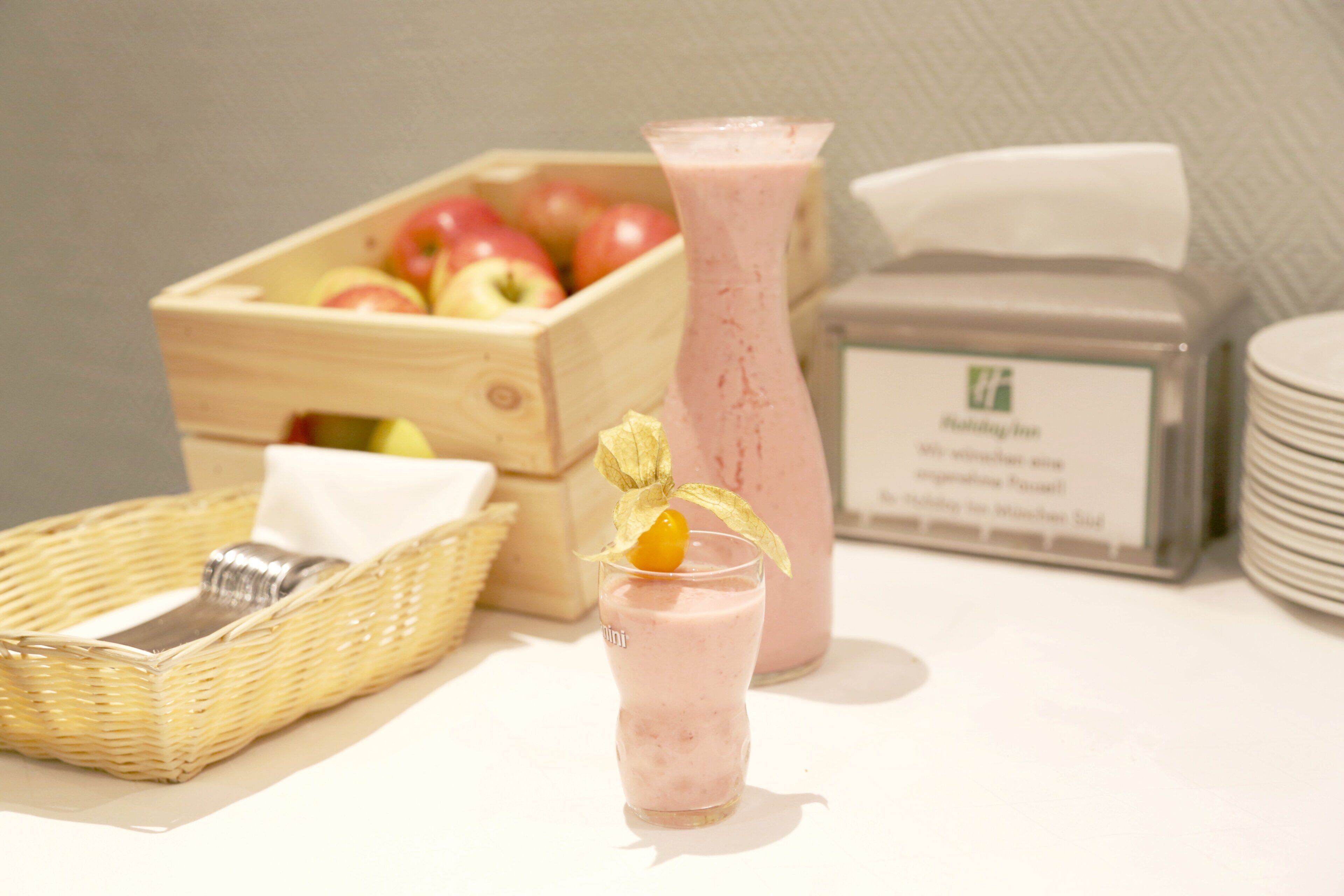 A wooden crate with apples, a glass of pink smoothie, a wicker basket, and a stack of white plates.