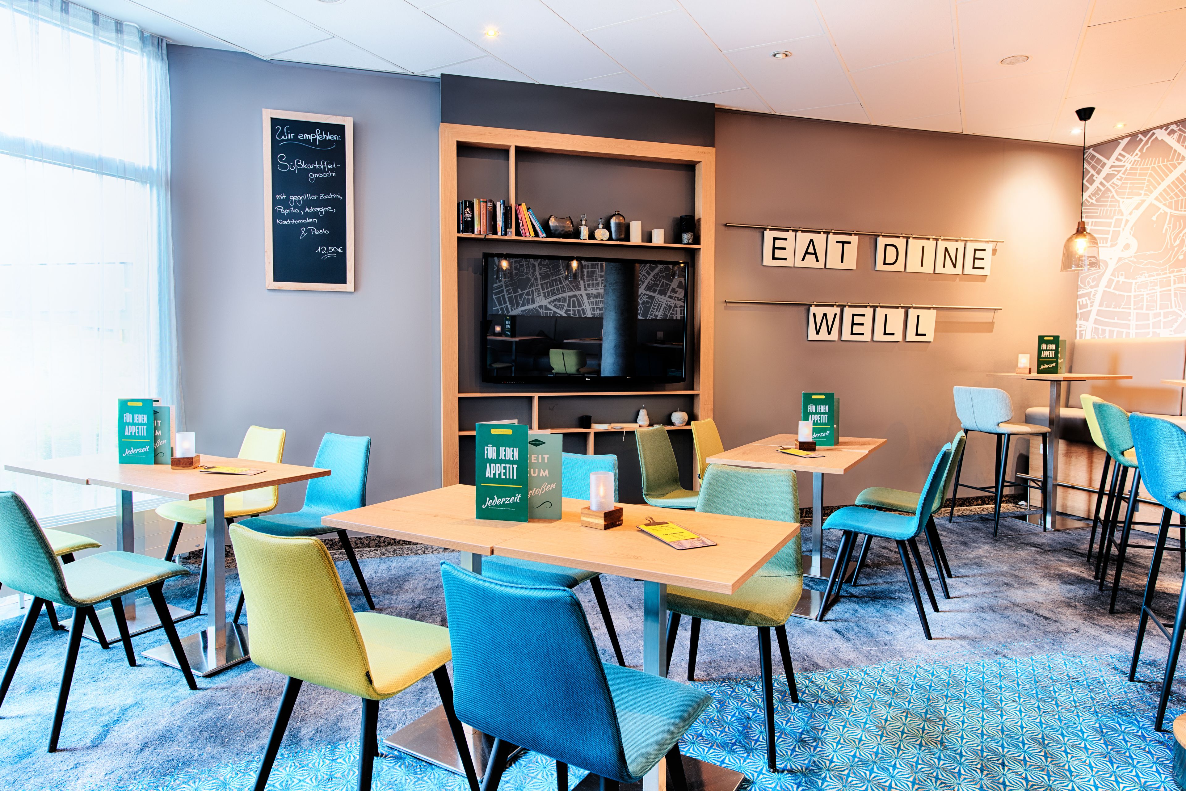 A modern dining area with tables and chairs, a bookshelf, and a chalkboard with writing.