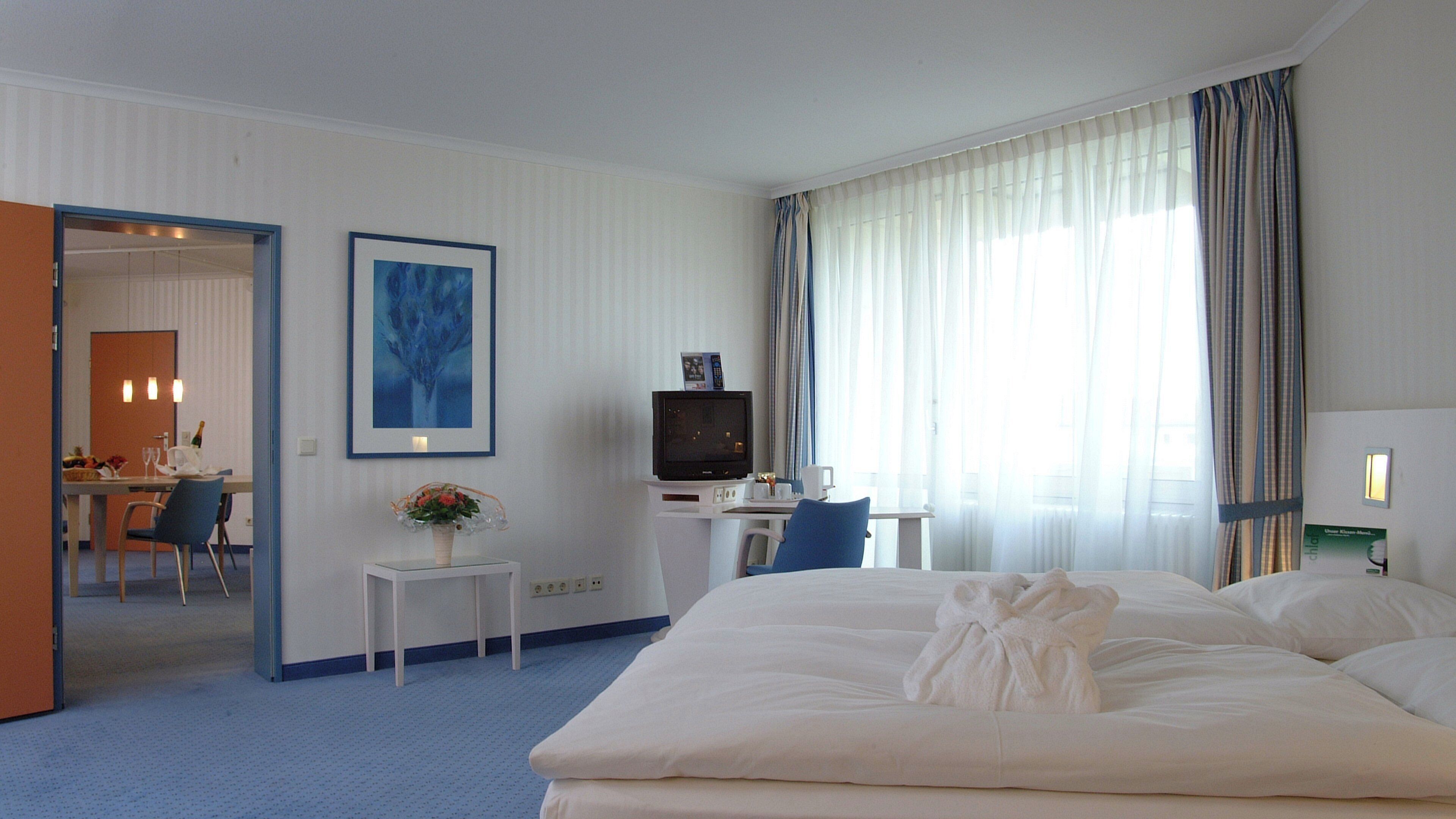 A hotel room with a bed, a desk with a television, a small table with a vase, and a dining area visible through an open door.