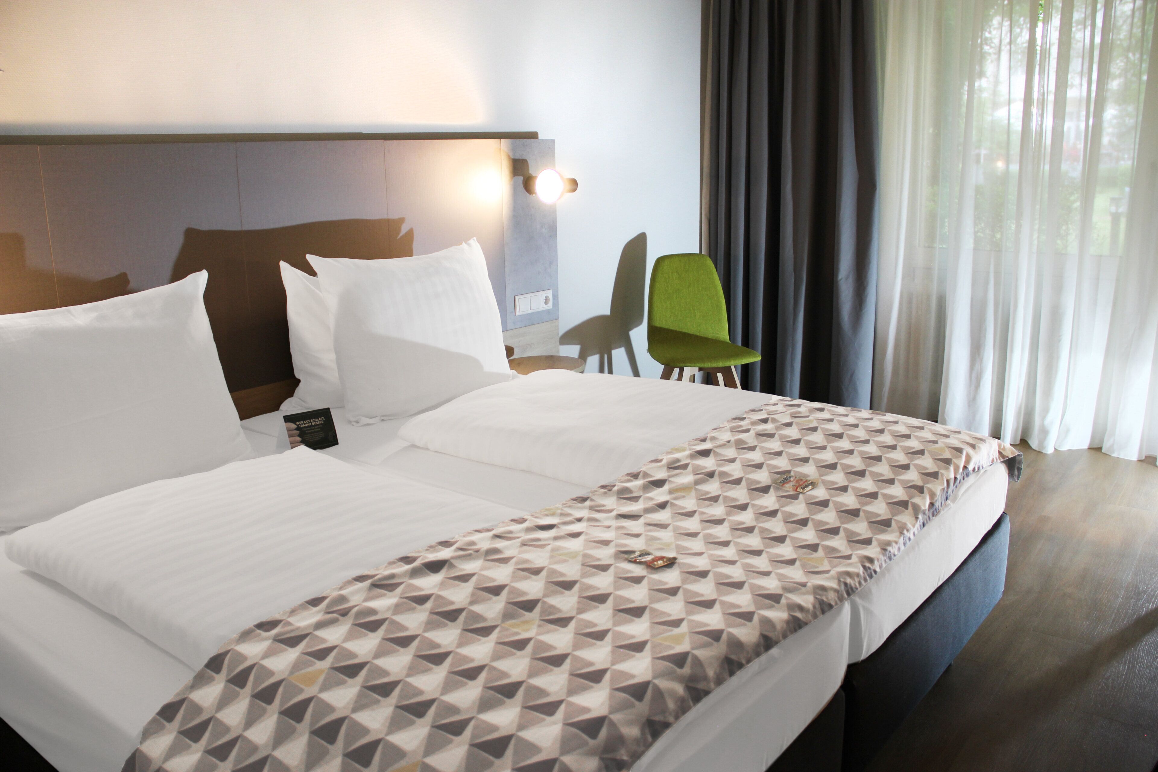 A hotel room with a bed, a headboard, a green chair, and a window with curtains.