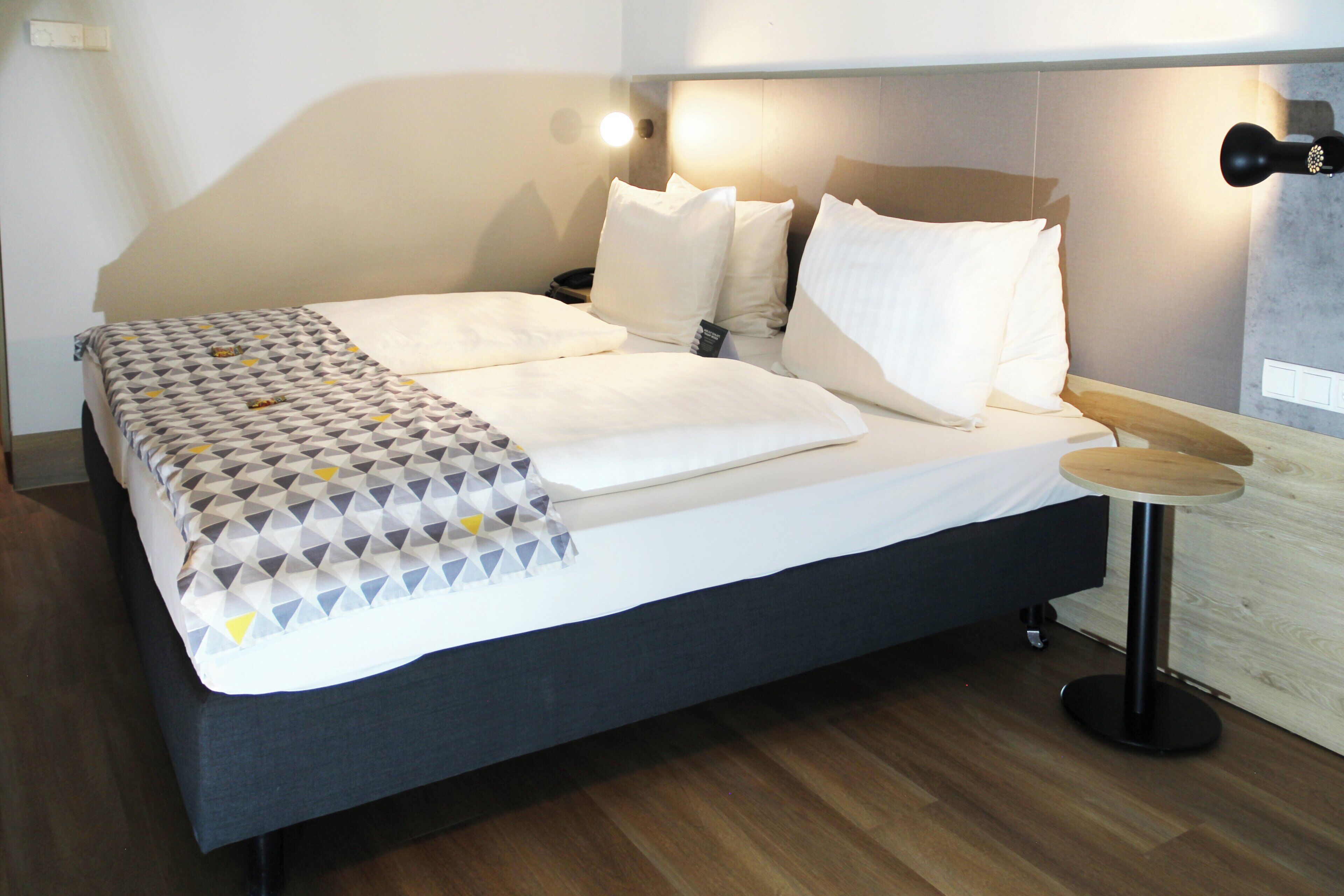 A double bed with a patterned blanket, a bedside table with a lamp, and a headboard with built-in lighting.