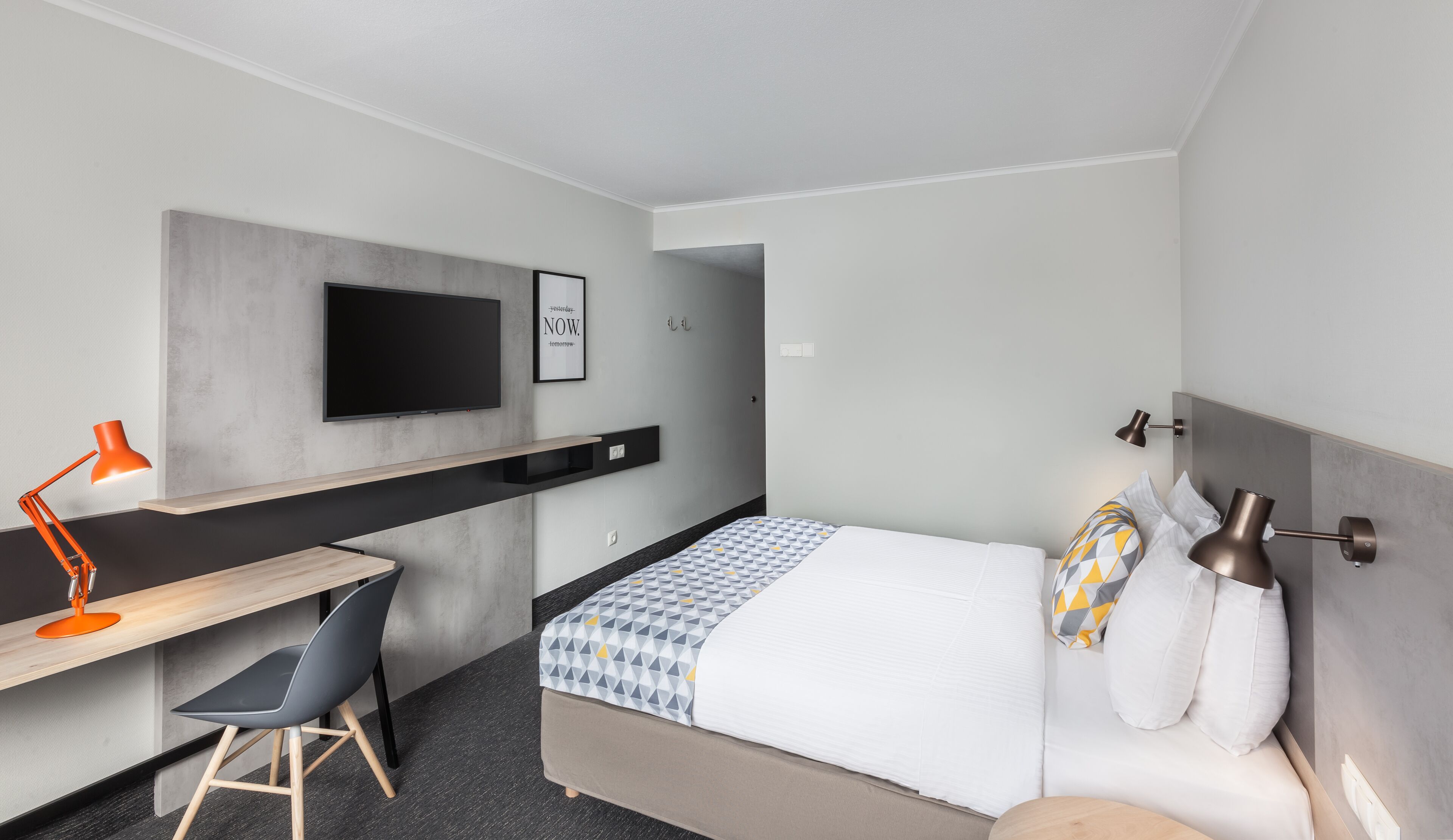 Una habitación de hotel moderna con una cama, un escritorio, una silla, un televisor y una lámpara de noche.