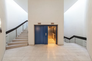 Un interior moderno con una puerta de ascensor azul, una escalera con barandillas de vidrio y un suelo de baldosas.