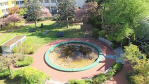 A circular water feature surrounded by a brick walkway, with trees and a building in the background. - Holiday Inn Munich - South by IHG (Munich)