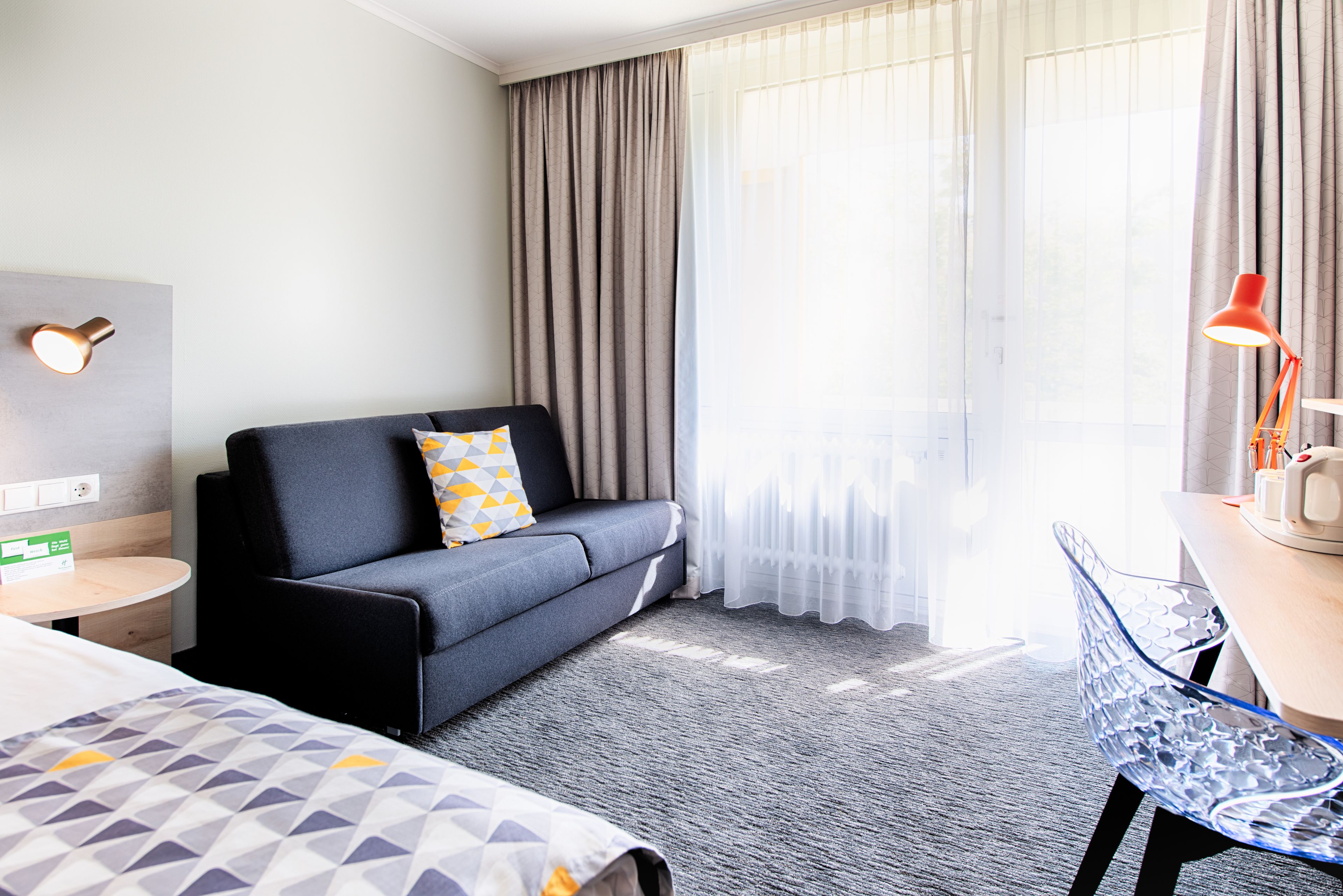 Ein Hotelzimmer mit grauem Sofa, einem kleinen runden Tisch, einem Bett mit gemusterten Bettbezügen und einem großen Fenster mit durchsichtigen Vorhängen.