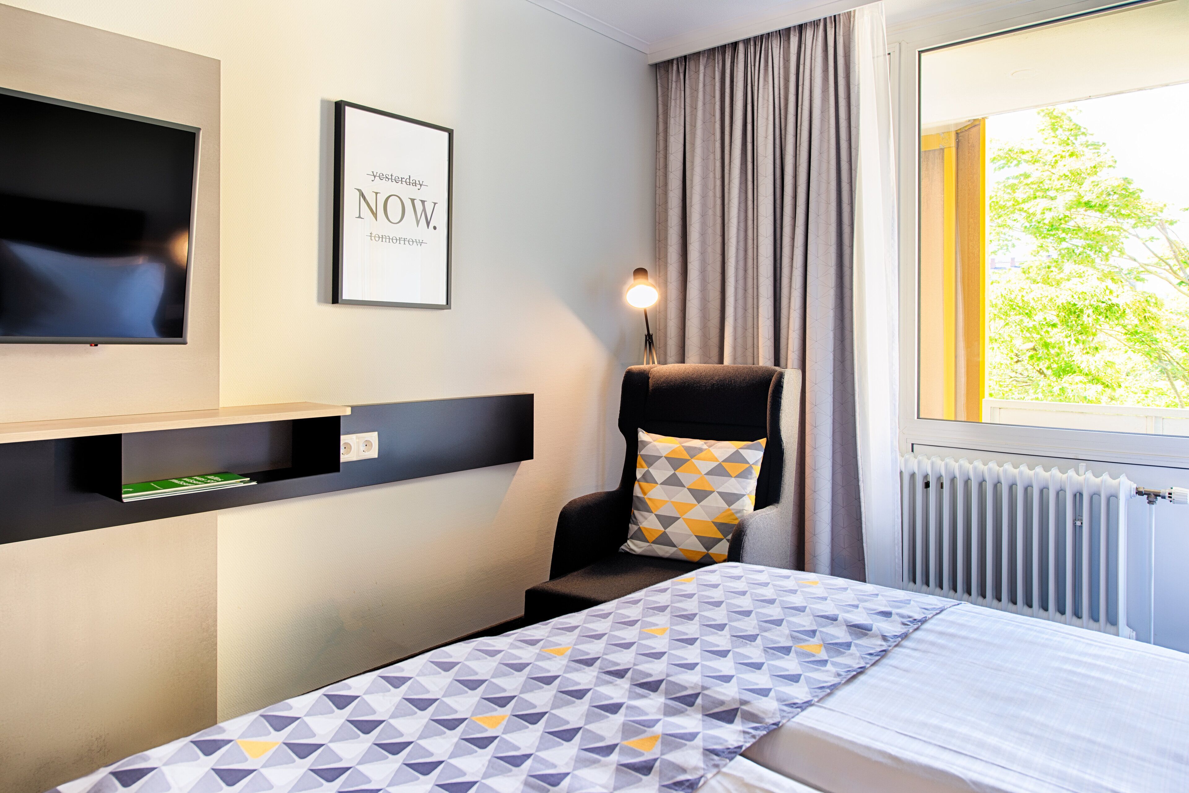 A hotel room with a bed, a chair, a TV, a framed quote on the wall, and a window with curtains.