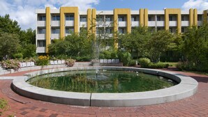 A building with a yellow facade, a circular pond with a fountain, and surrounding greenery. - Holiday Inn Munich - South by IHG (Munich)