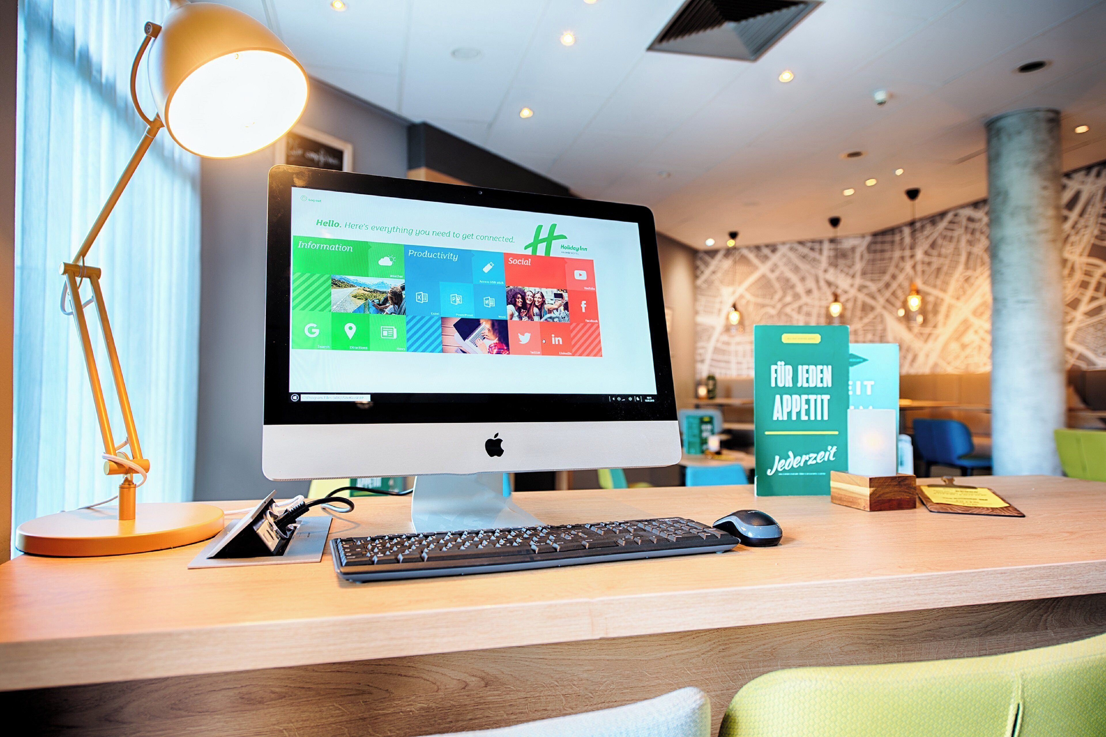 Una computadora de escritorio con un monitor que muestra la interfaz de una aplicación de hotel, un teclado, un ratón y una lámpara sobre un escritorio de madera.